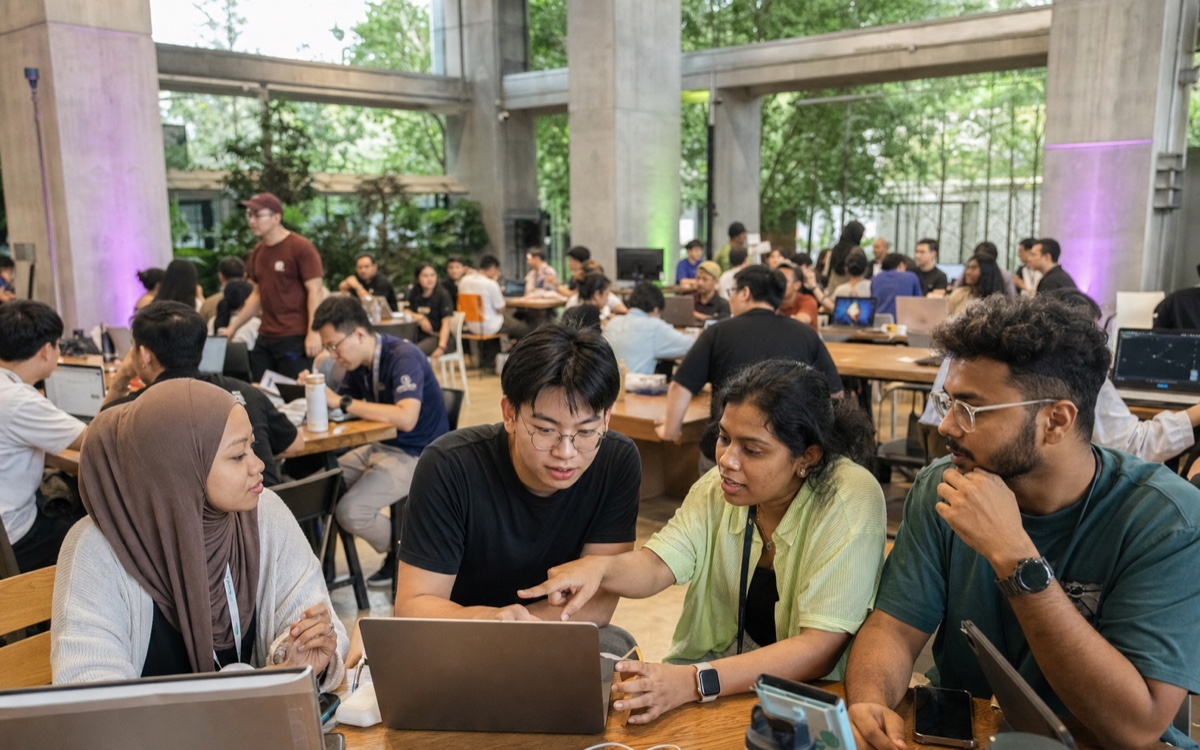 Long shared tables full of people building on laptops in a plant-filled hall